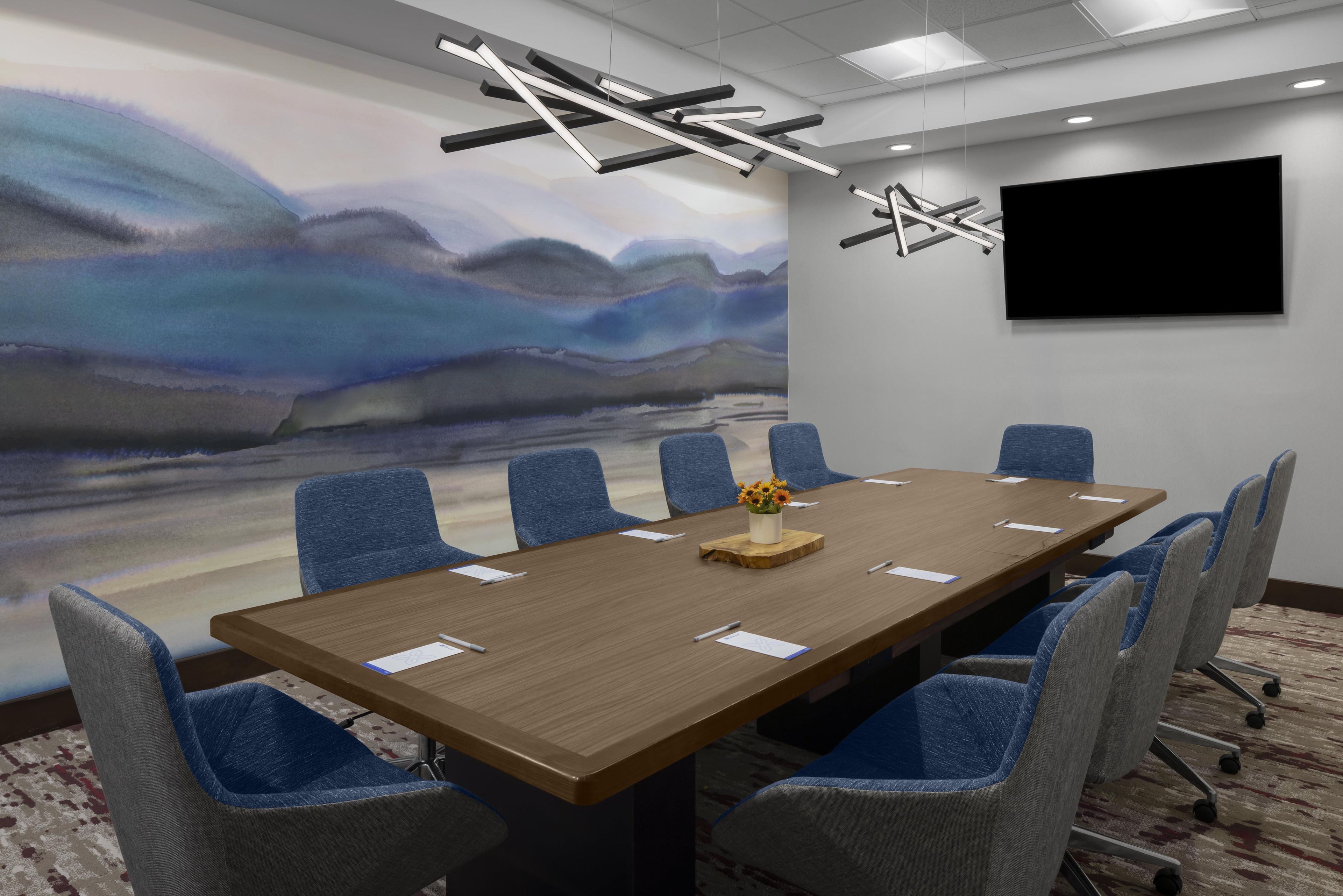 Un salón de conferencias con una larga mesa, sillas azules, un cuadro, un televisor de pantalla plana y una lámpara moderna.