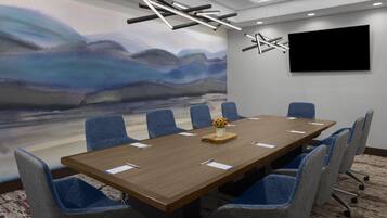 Un salón de conferencias con una larga mesa, sillas azules, un cuadro, un televisor de pantalla plana y una lámpara moderna.