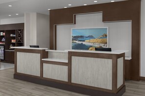 A reception desk with a framed landscape painting, a wooden reception desk, and a shelving unit with various items. - Hilton Garden Inn San Marcos (San Marcos)