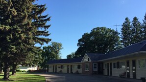 A motel with multiple rooms, a covered parking area, and a large tree in the foreground. - Yellow Quill Motel (Portage La Prairie)