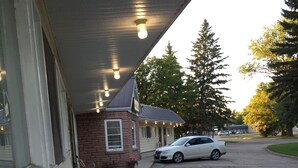 A building with a porch, a white car parked in front, and a potted plant with flowers. - Yellow Quill Motel (Portage La Prairie)