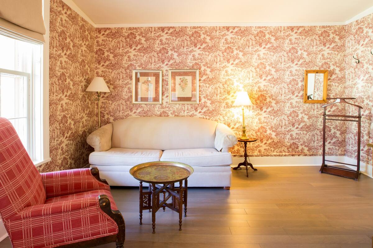 A living room with a white sofa, a plaid armchair, a small round table, and a lamp. There are two framed pictures on the wall and a mirror on a stand.