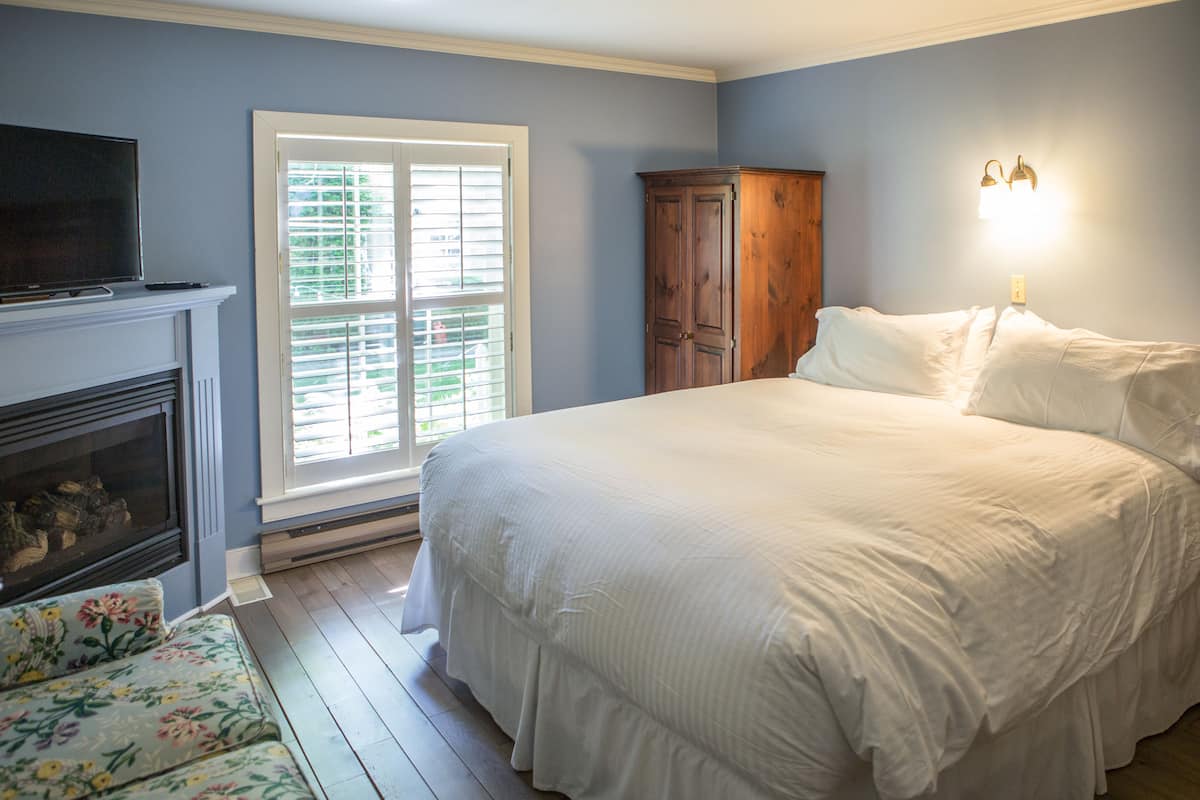 A bedroom with a large bed, a fireplace, a window with blinds, and a wooden wardrobe.
