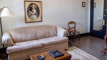 Uma sala de estar com um sofá bege, uma mesa de centro de madeira com um controle remoto e um livro, uma cadeira de madeira, um retrato emoldurado na parede e uma porta escura com um espelho.