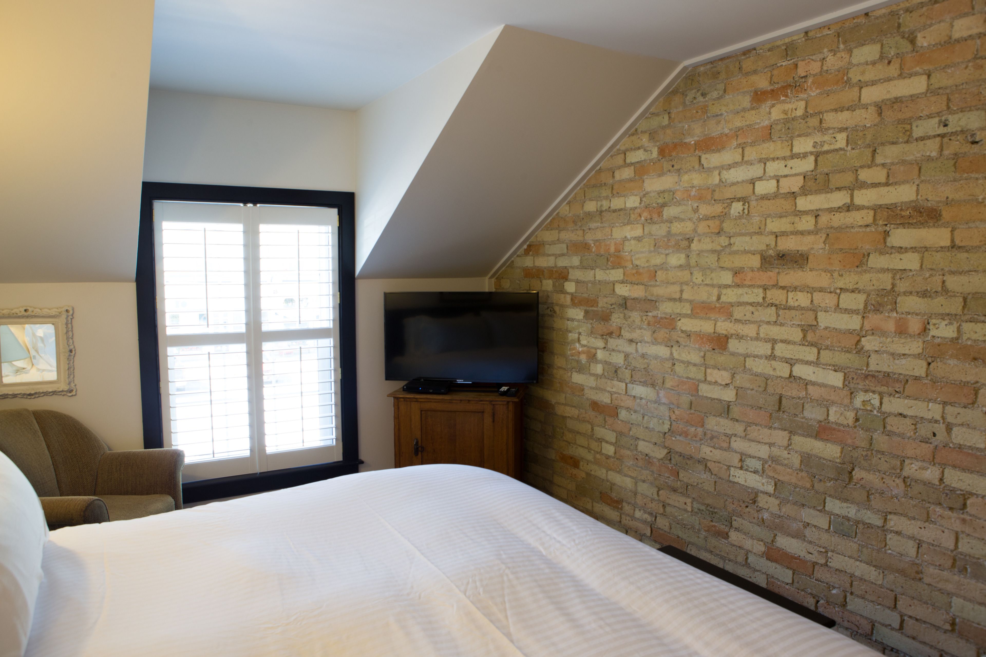 Un dormitorio con una cama grande, un televisor sobre un mueble de madera, una pared de ladrillo y una ventana con persianas.