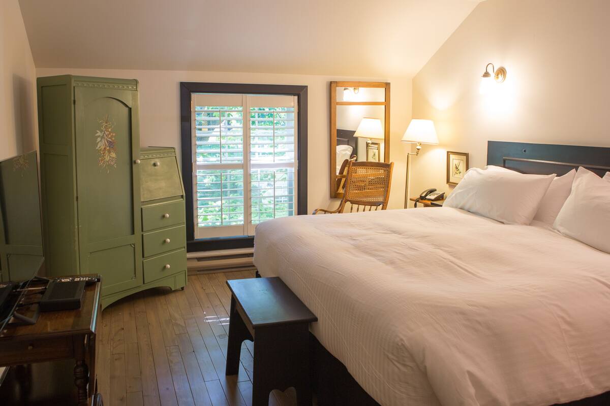 A hotel room with a large bed, a wooden desk, a green dresser, a chair, and a window with a view of greenery.