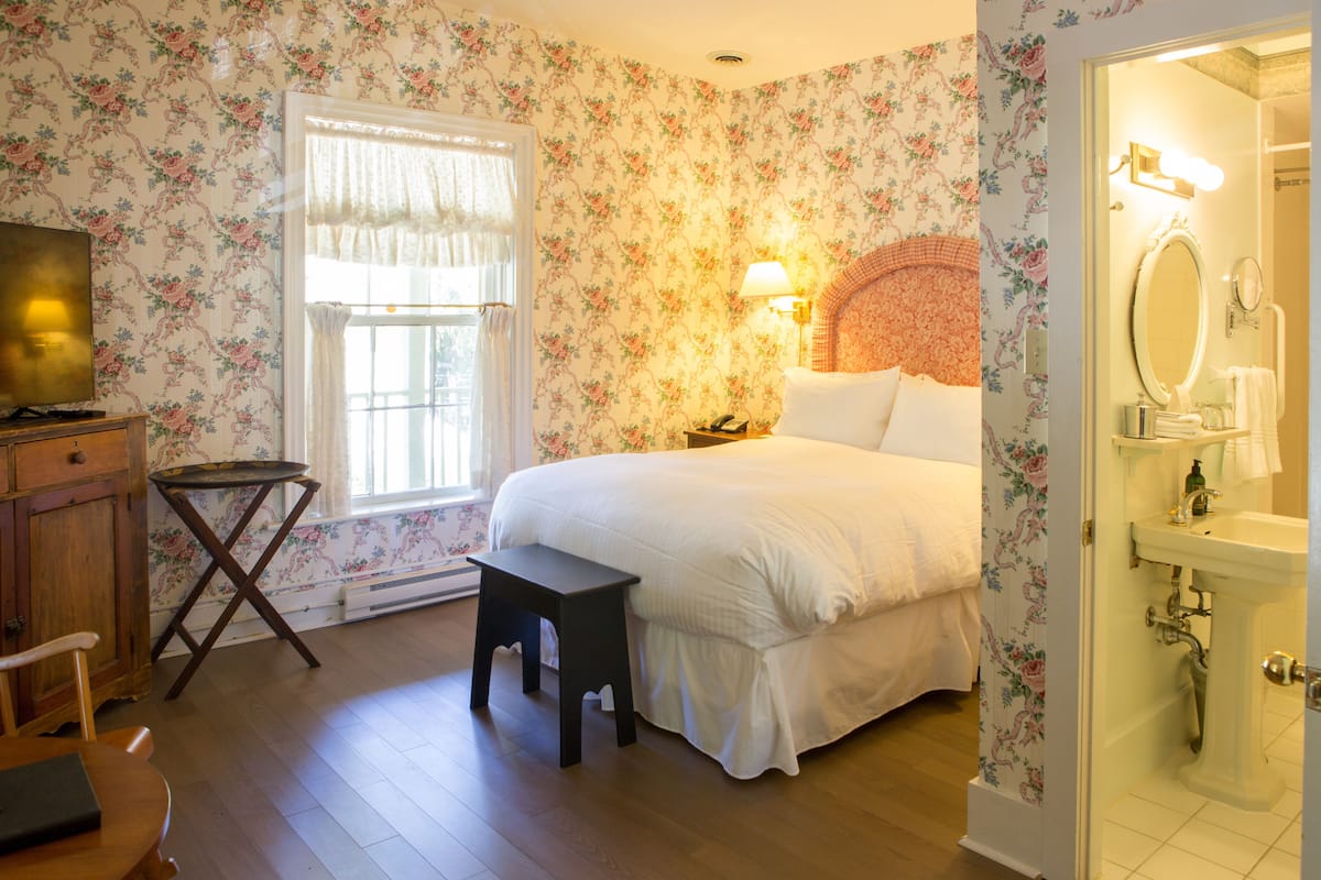 A bedroom with floral wallpaper, a bed, a nightstand, a lamp, a dresser, and a television.