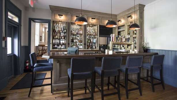 A bar area with wooden bar stools, a bar counter, and shelves stocked with bottles. - The Little Inn of Bayfield (Bluewater)