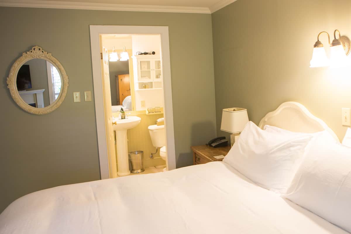 A hotel room with a bed, a nightstand with a lamp, a mirror, and a bathroom visible through an open door.