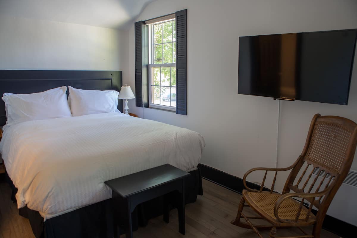 A neatly made bed with white linens, a bedside table with a lamp, a wooden rocking chair, and a flat-screen TV mounted on the wall.