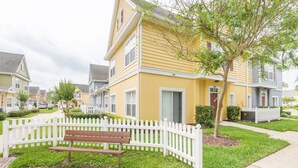 A row of yellow townhouses with white picket fences, a bench, and a tree in the foreground. - Villas at Seven Dwarf by FunQuest (Kissimmee)