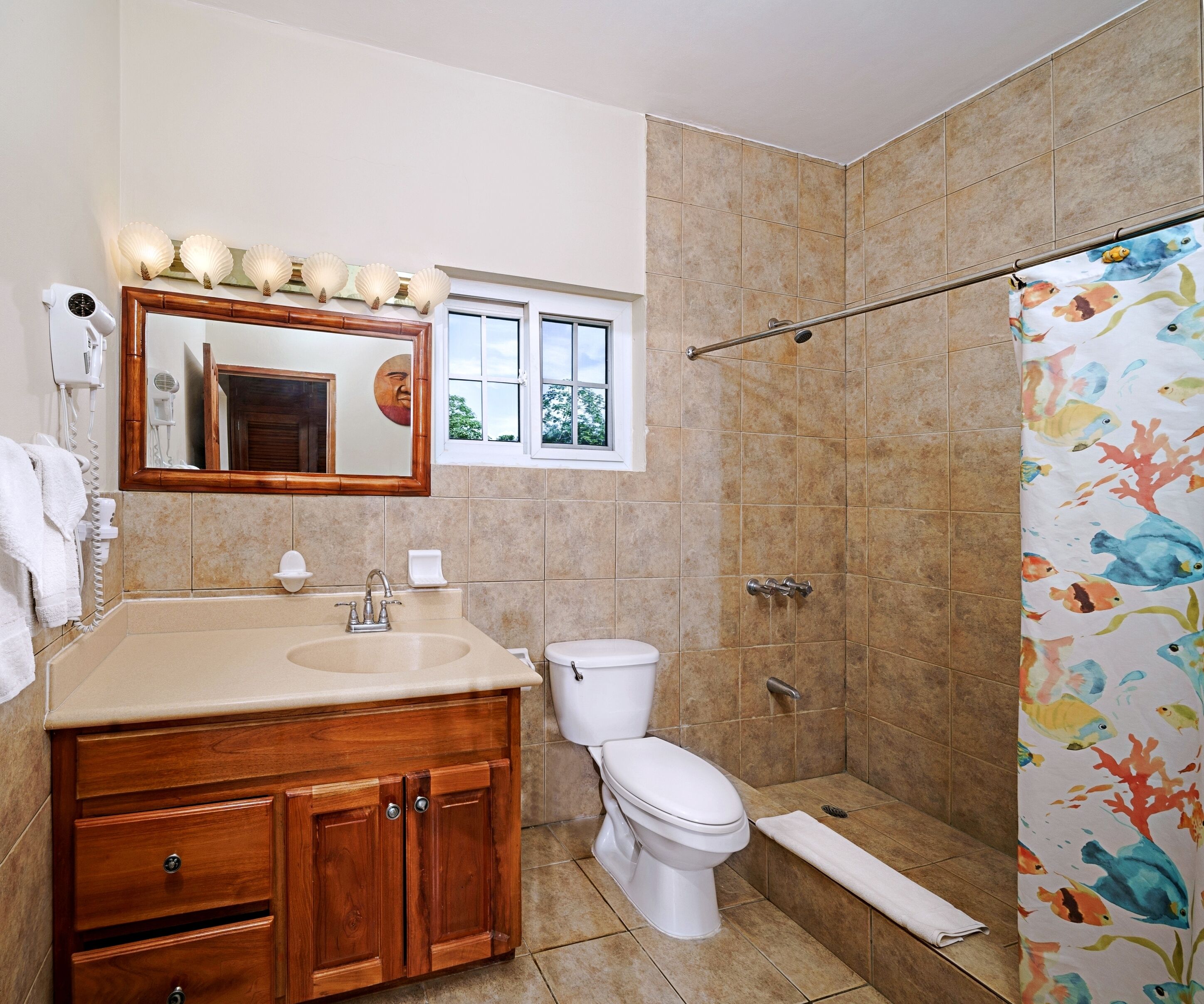 A bathroom with a wooden vanity, a toilet, a shower with a fish-themed curtain, and a window with a view of trees.