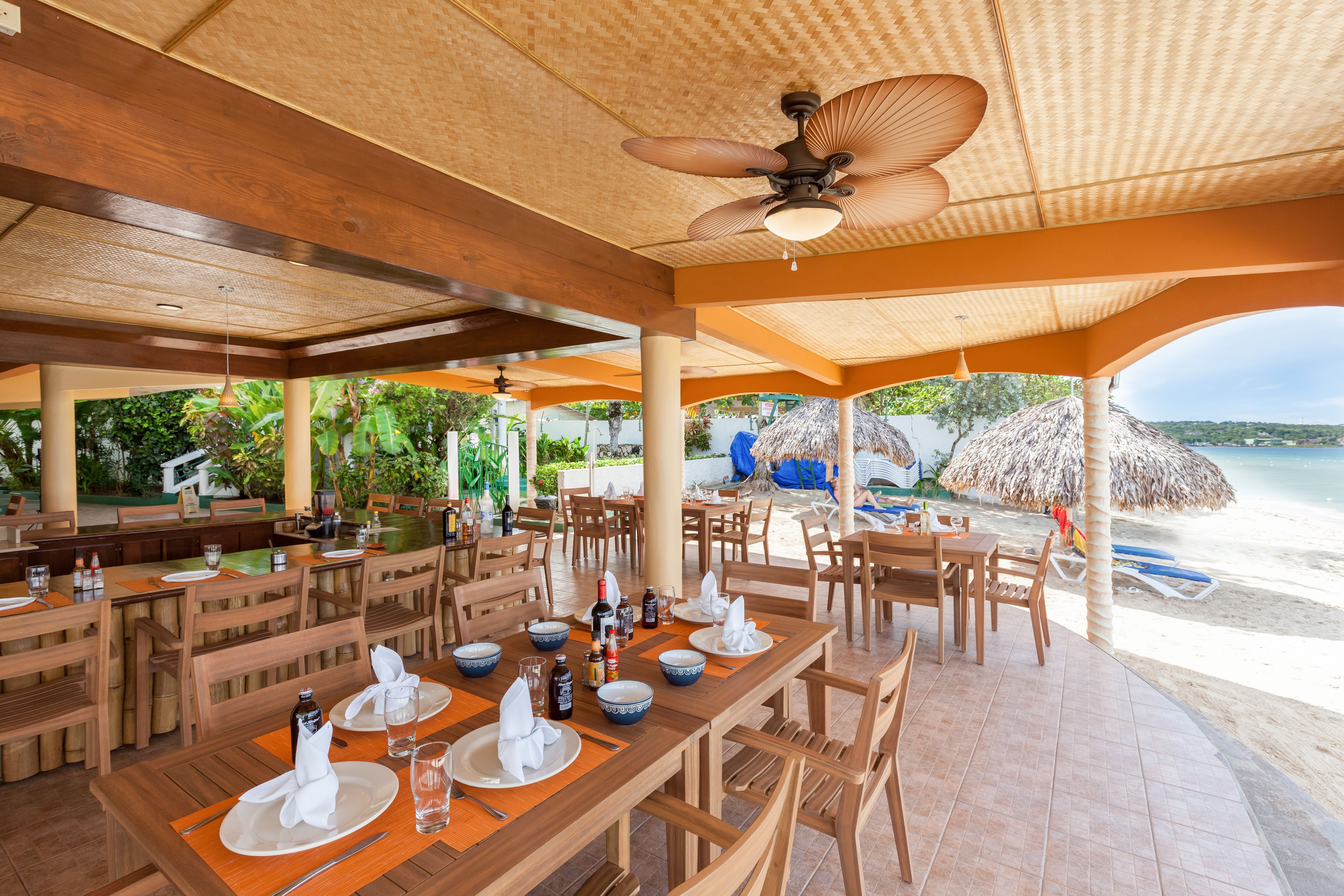 A restaurant with wooden tables and chairs, a ceiling fan, and a view of the beach.