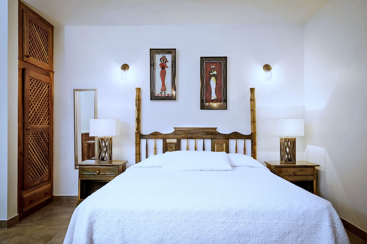Un dormitorio ordenado con una cama grande, cabecera de madera, dos mesitas de noche con lámparas, un espejo y dos cuadros enmarcados en la pared.