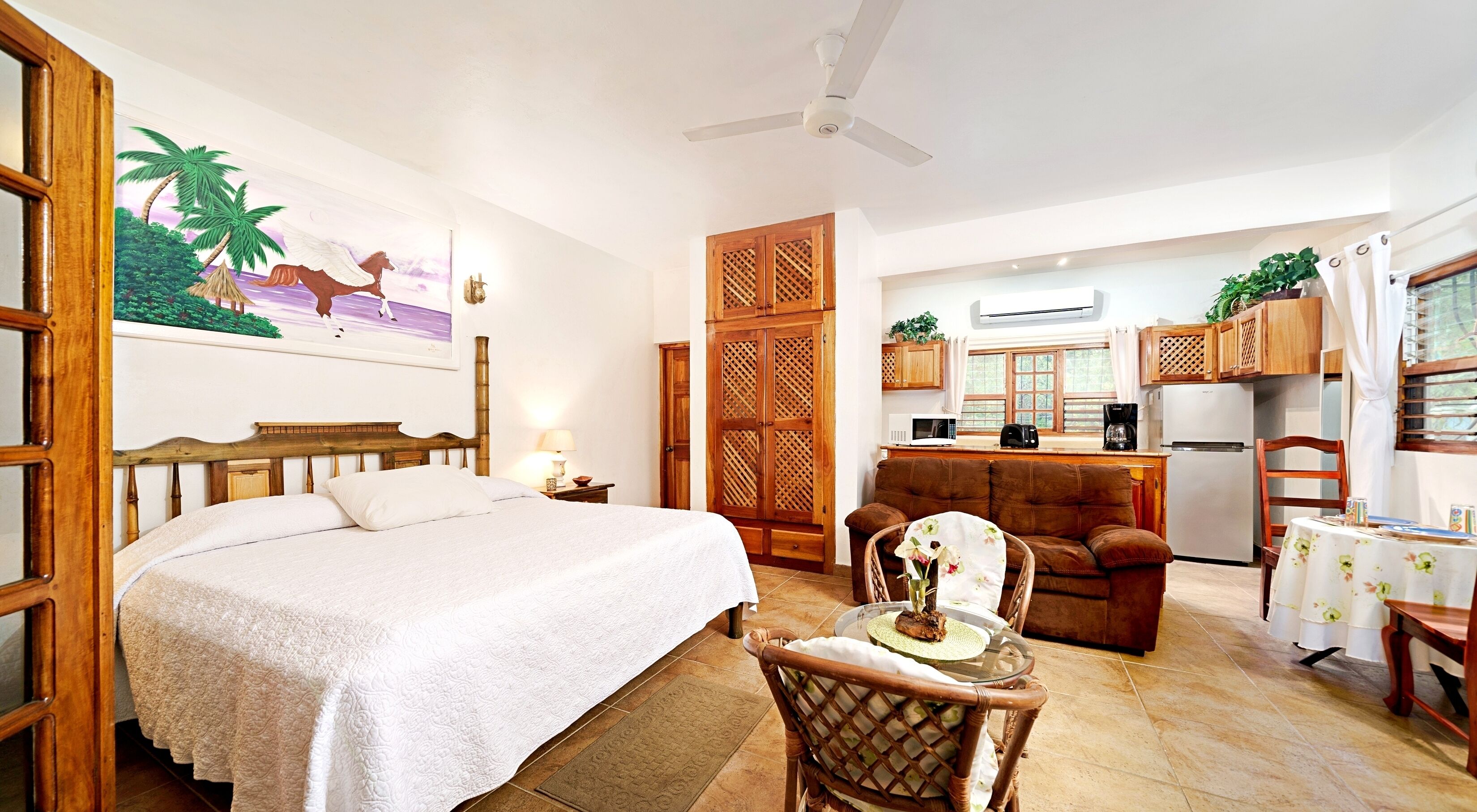 A bedroom with a large bed, a wooden wardrobe, a dining area with a table and chairs, and a kitchen with a refrigerator and microwave.