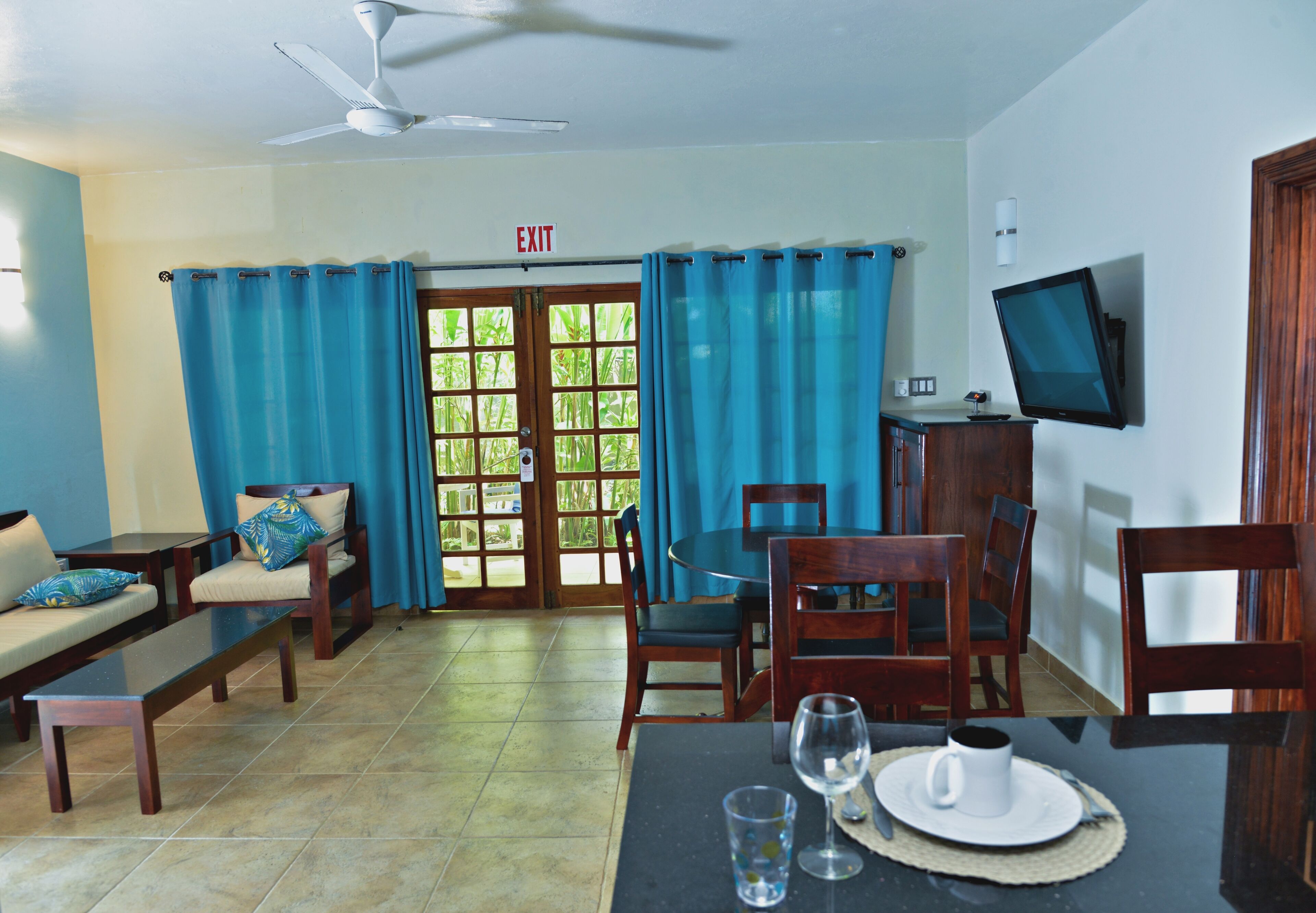A dining area with a table set for two, a television mounted on the wall, and a view of the outdoors through a door with curtains.