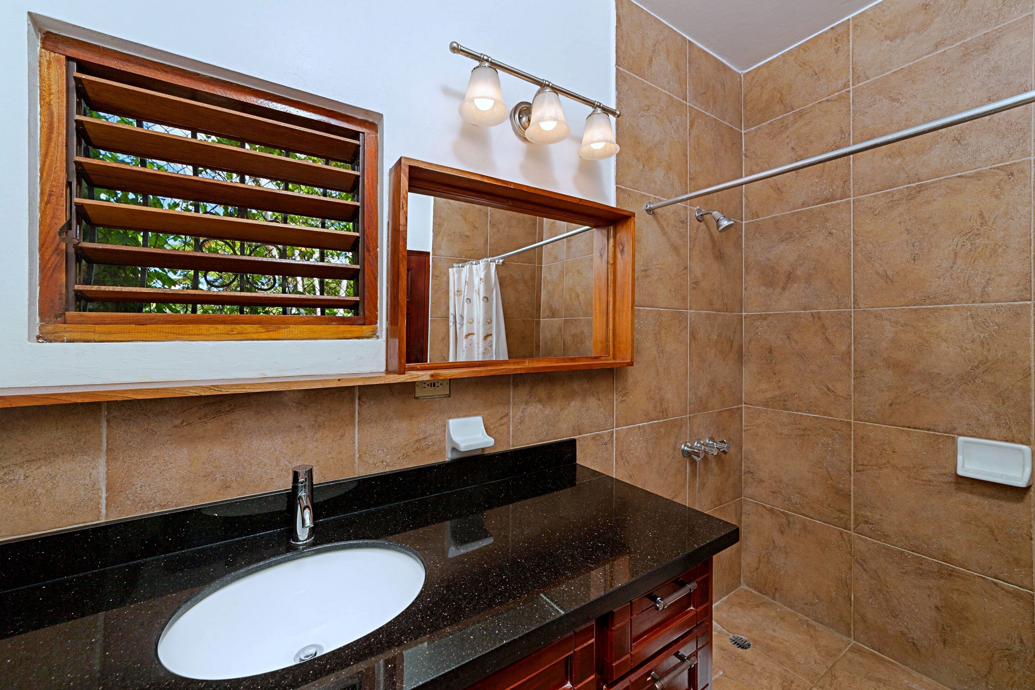 Un baño con lavamanos de granito negro, ventana con marco de madera y persianas, espejo con iluminación y paredes de azulejos.