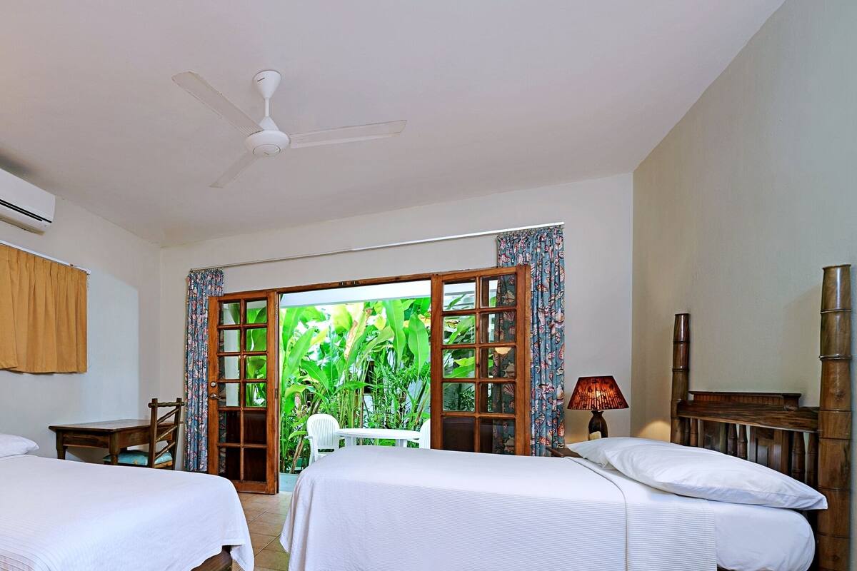 Habitación de hotel con dos camas, ventilador de techo, aire acondicionado y vista a la vegetación a través de una puerta abierta.