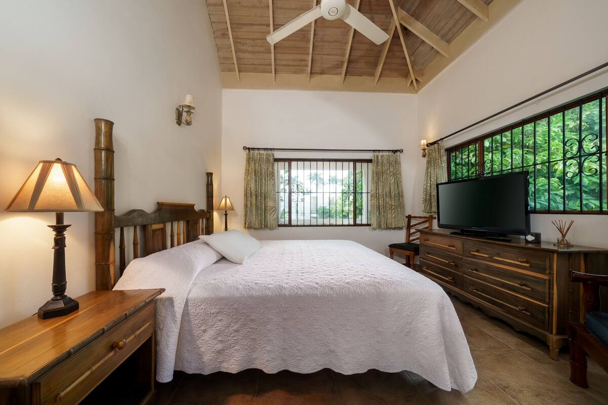 Un dormitorio con cama, televisor, cómoda de madera, lámpara y ventana con cortinas.