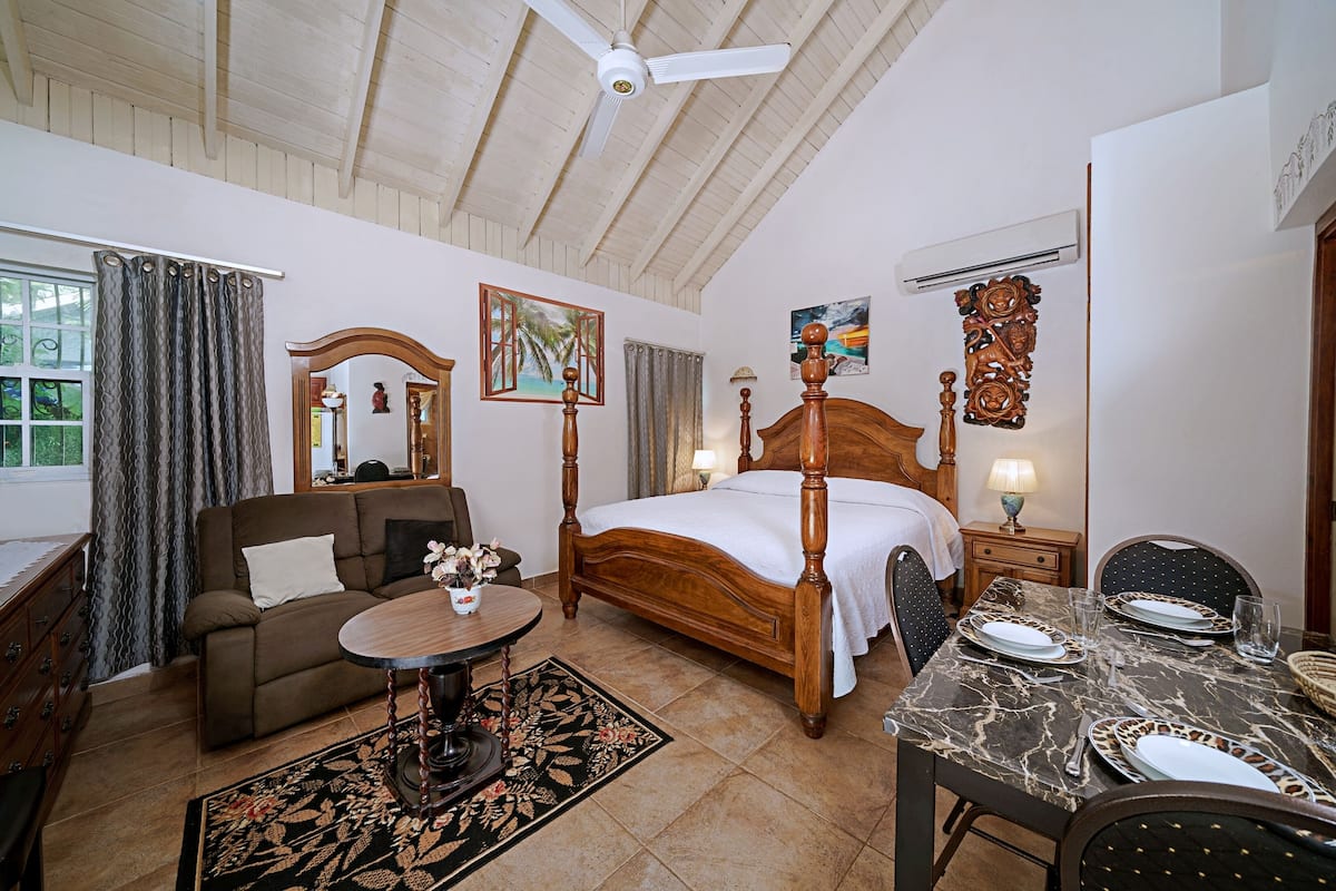 Un dormitorio con cama de madera, zona de estar con sofá, mesa de comedor para dos personas y ventilador de techo.