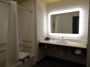 Une salle de bain avec un lavabo blanc, un miroir doté d’un éclairage intégré, un rideau de douche et un porte-serviettes.