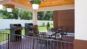 Un patio exterior con techo de madera, dos parrillas de barbacoa, una mesa con sillas y cestas de flores colgantes.