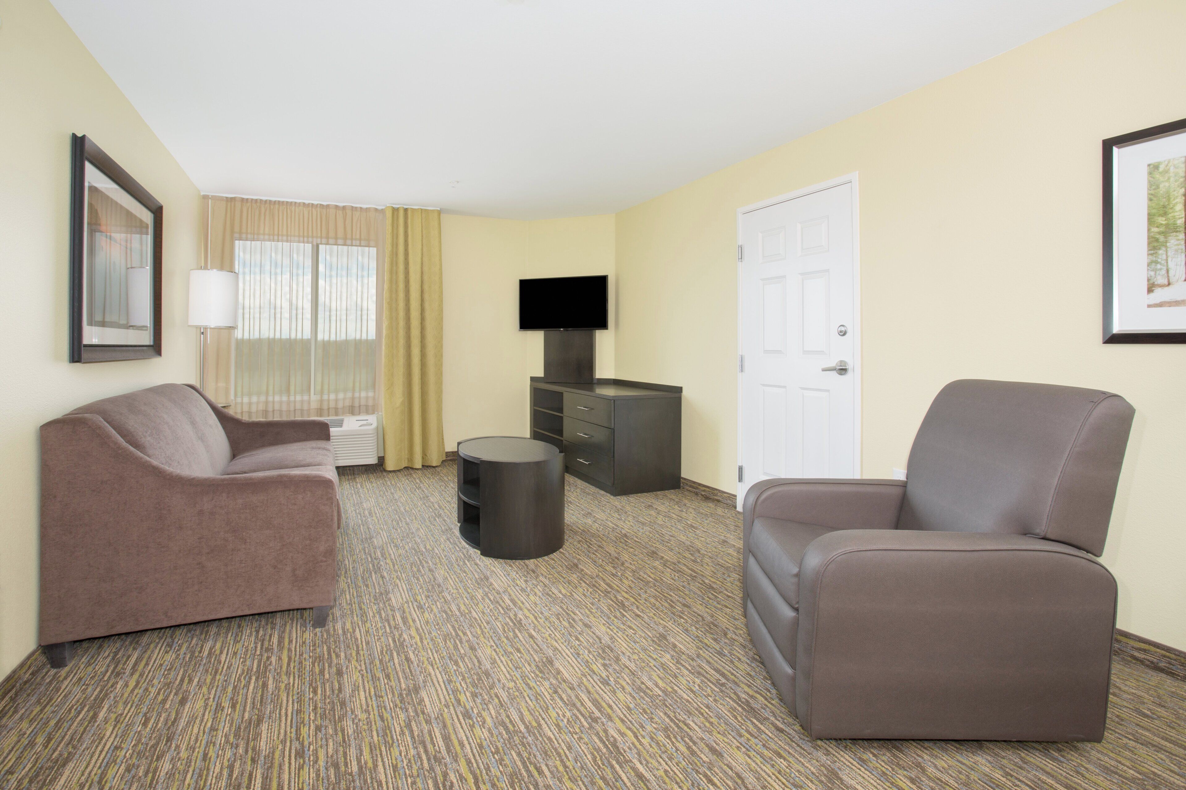 A hotel room with two sofas, a flat-screen TV, a small round table, a dresser, and a door.