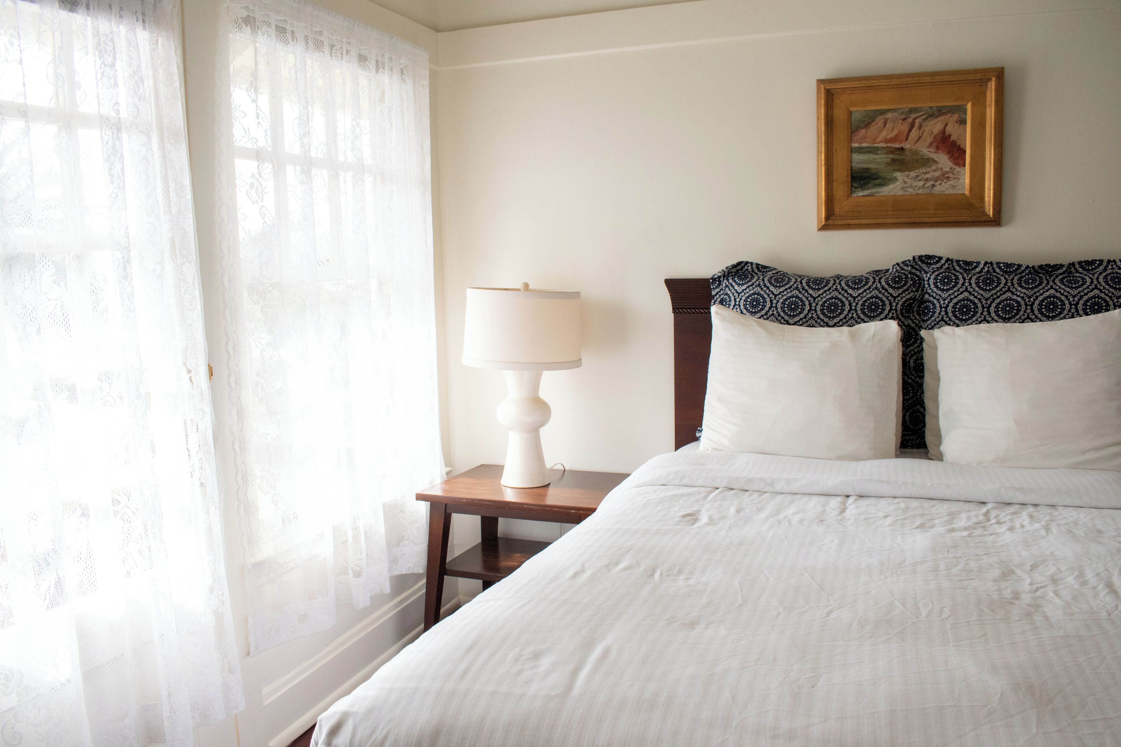 A neatly made bed with white linens, a wooden nightstand with a white lamp, and a framed painting on the wall.