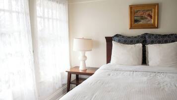 A neatly made bed with white linens, a wooden nightstand with a white lamp, and a framed painting on the wall.