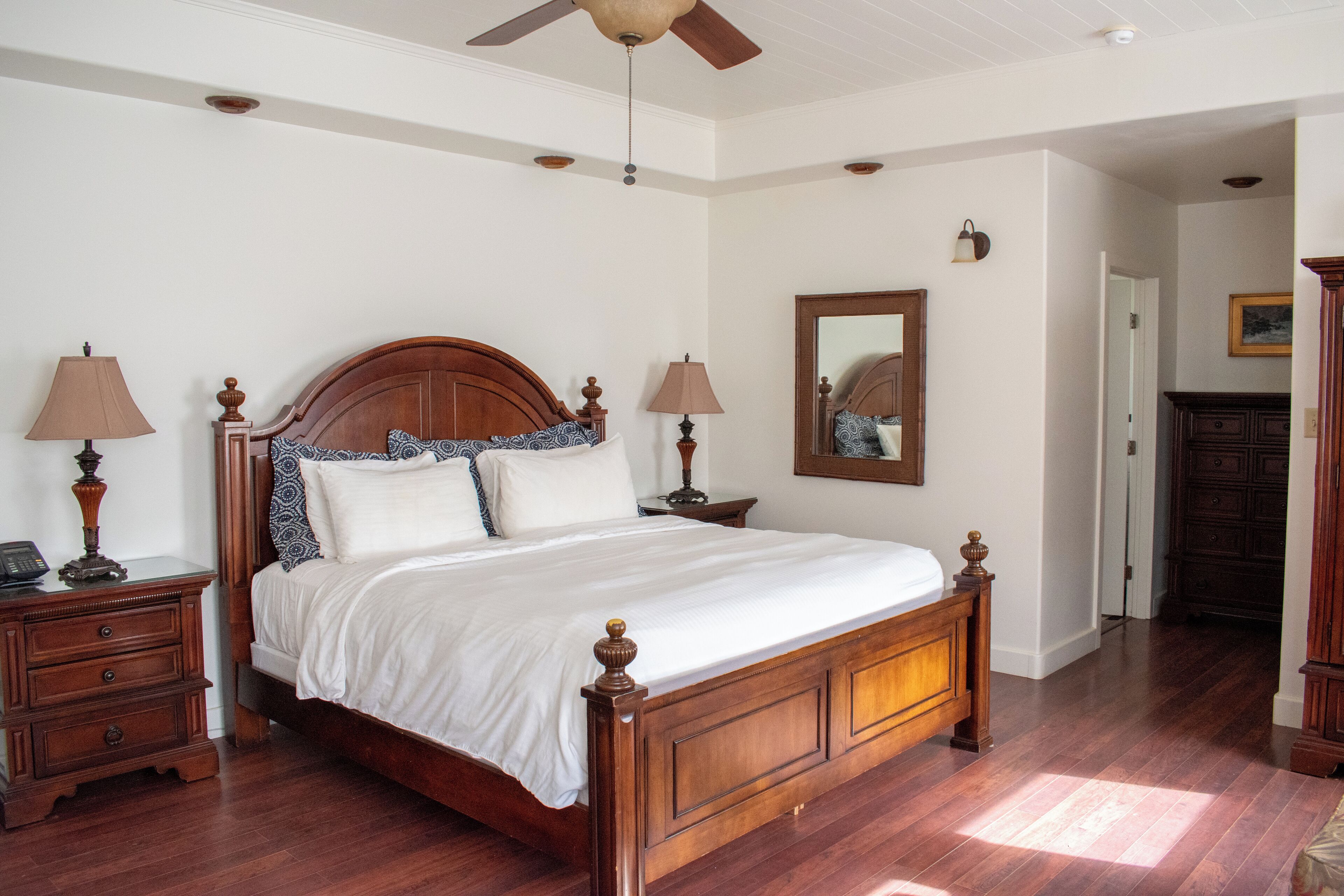 A bedroom with a wooden bed, two bedside tables with lamps, a ceiling fan, a mirror on the wall, and a wooden floor.