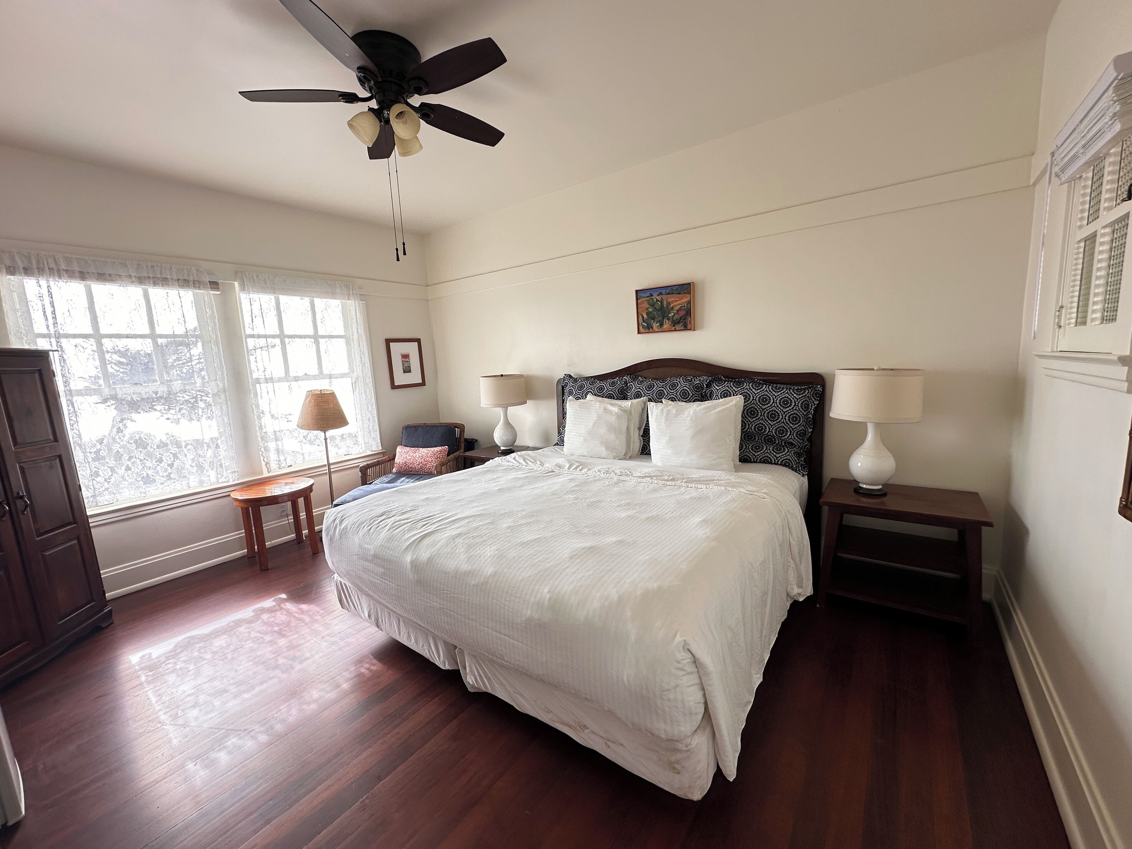 A bedroom with a large bed, two bedside tables with lamps, a ceiling fan, a window with sheer curtains, and a wooden floor.