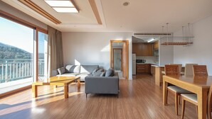 A modern living room with a sofa, coffee table, and dining area. There is a large window with a view of a balcony and a door leading to a kitchen. - Elysian Gangchon Resort (Chuncheon)