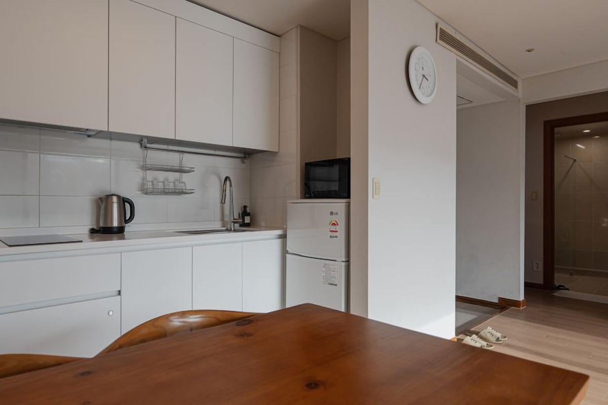 A modern kitchen with white cabinets, a stainless steel kettle, a microwave, and a clock on the wall.