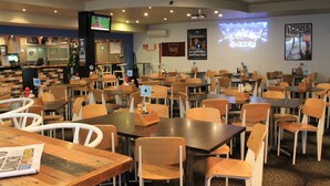 A restaurant with multiple tables and chairs, a bar area, and a television screen displaying a sports game. - Hotel Beach House Nambour (Nambour)