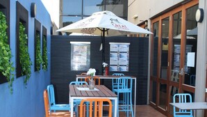 An outdoor patio with wooden tables and blue chairs, a white umbrella, and a black wall with green plants. - Hotel Beach House Nambour (Nambour)