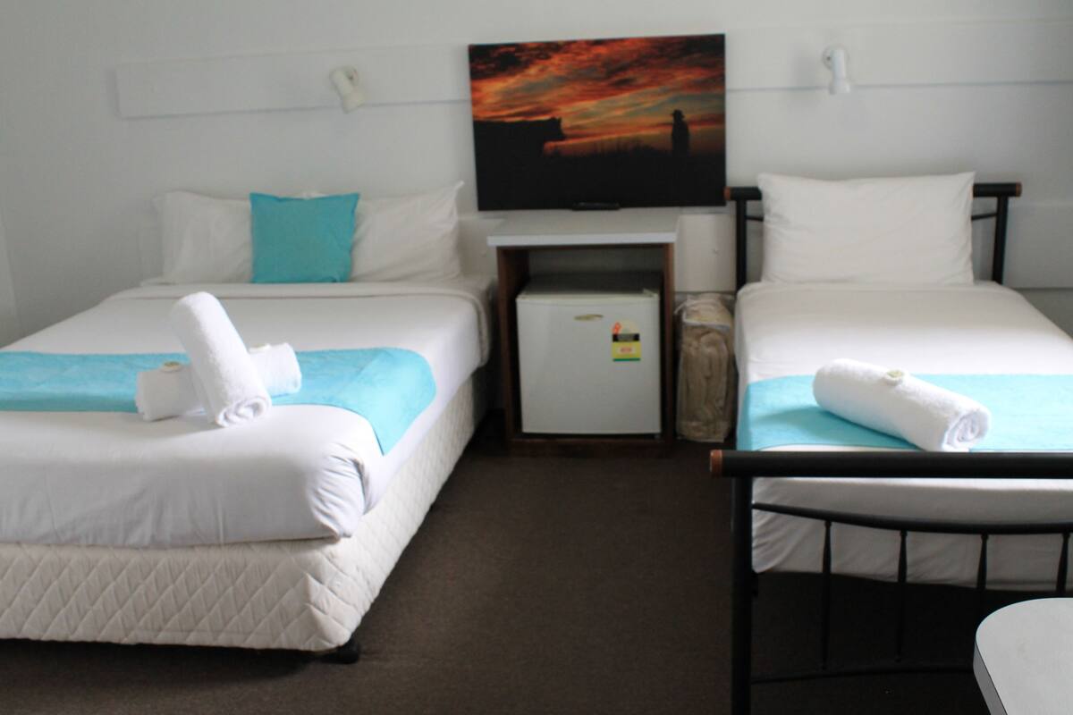 Habitación de hotel con dos camas, cada una con un colchón blanco y una manta azul. Hay un refrigerador pequeño y un televisor en la pared.