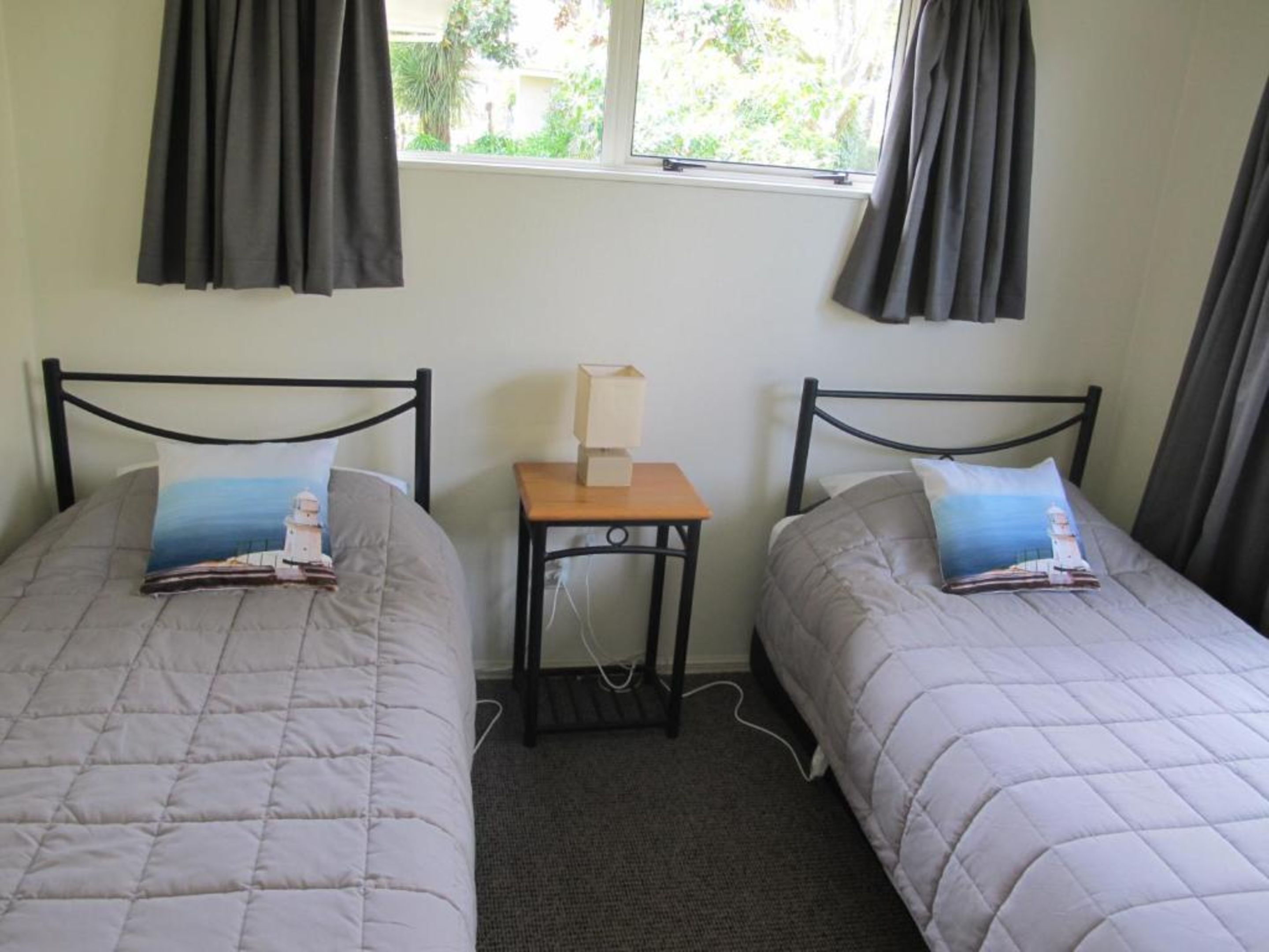 A room with two single beds, each with a pillow featuring a lighthouse design. There is a small wooden nightstand between the beds with a lamp on it. The room has a window with curtains, and the walls are plain.