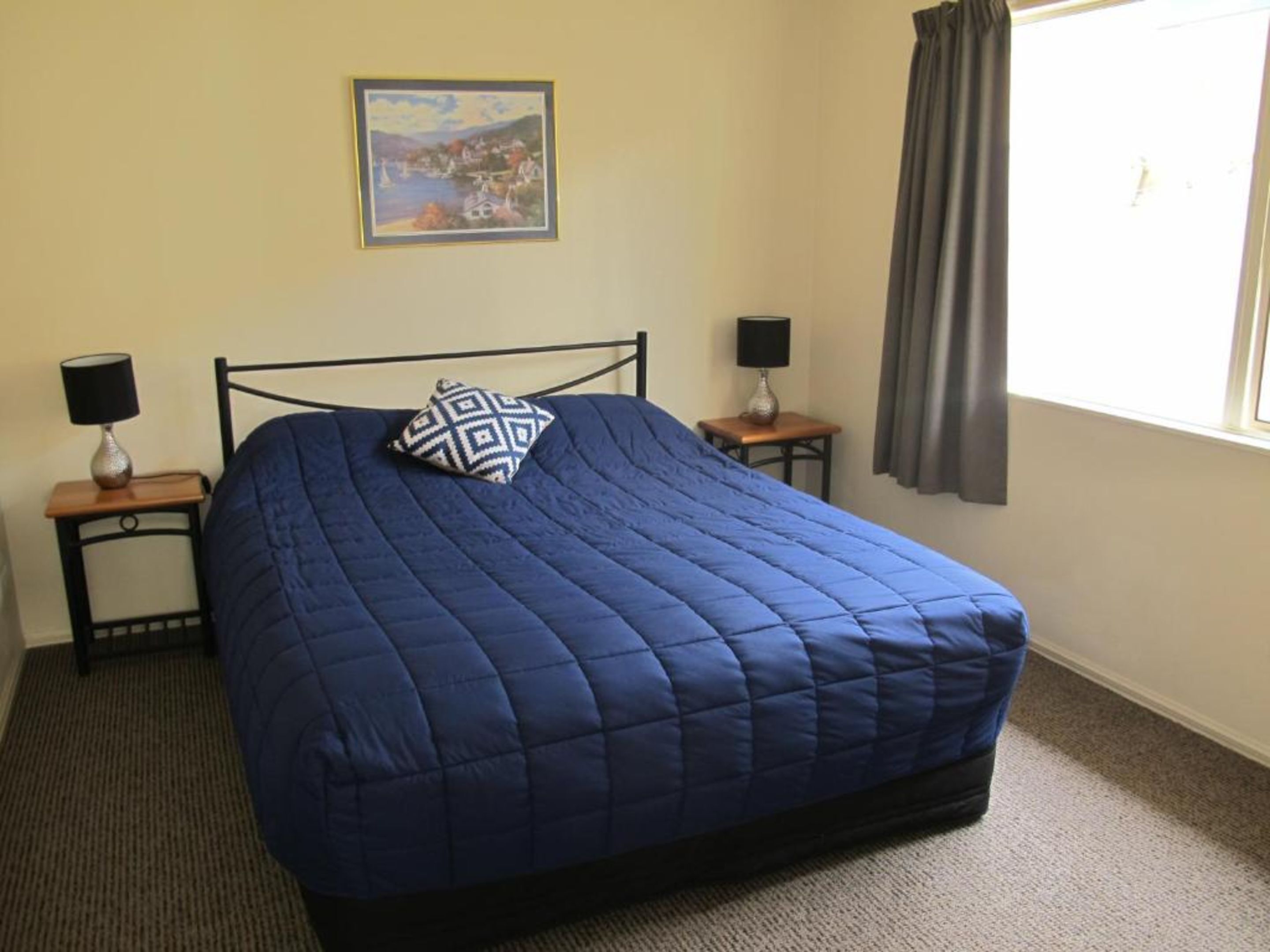 A neatly made bed with a blue quilt, two bedside tables with lamps, a painting on the wall, and a window with curtains.