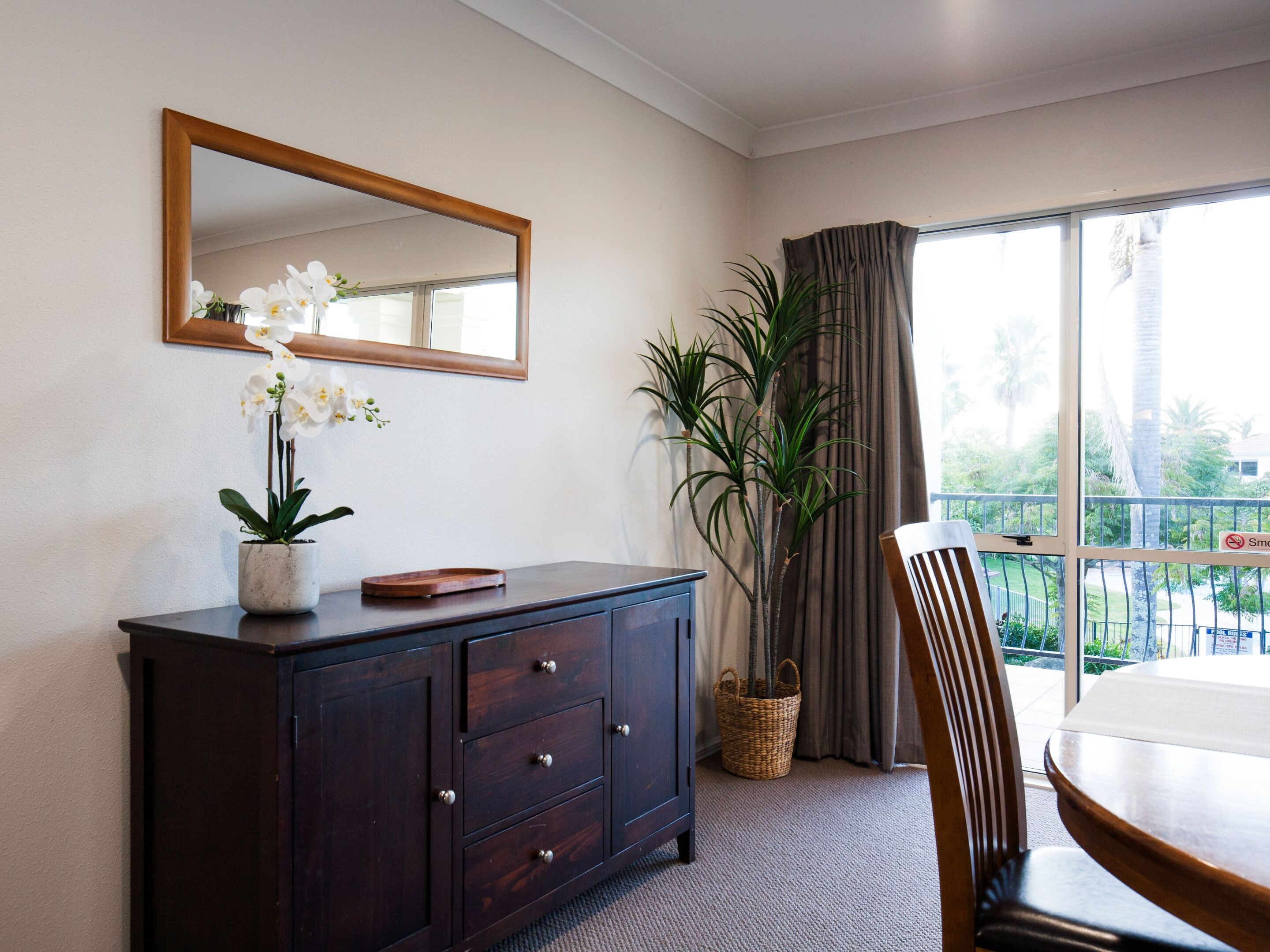 Una habitación con un armario de madera oscura, un espejo, una orquídea en maceta, una planta alta en una cesta, una mesa de comedor y vistas a palmeras a través de una ventana con cortinas.