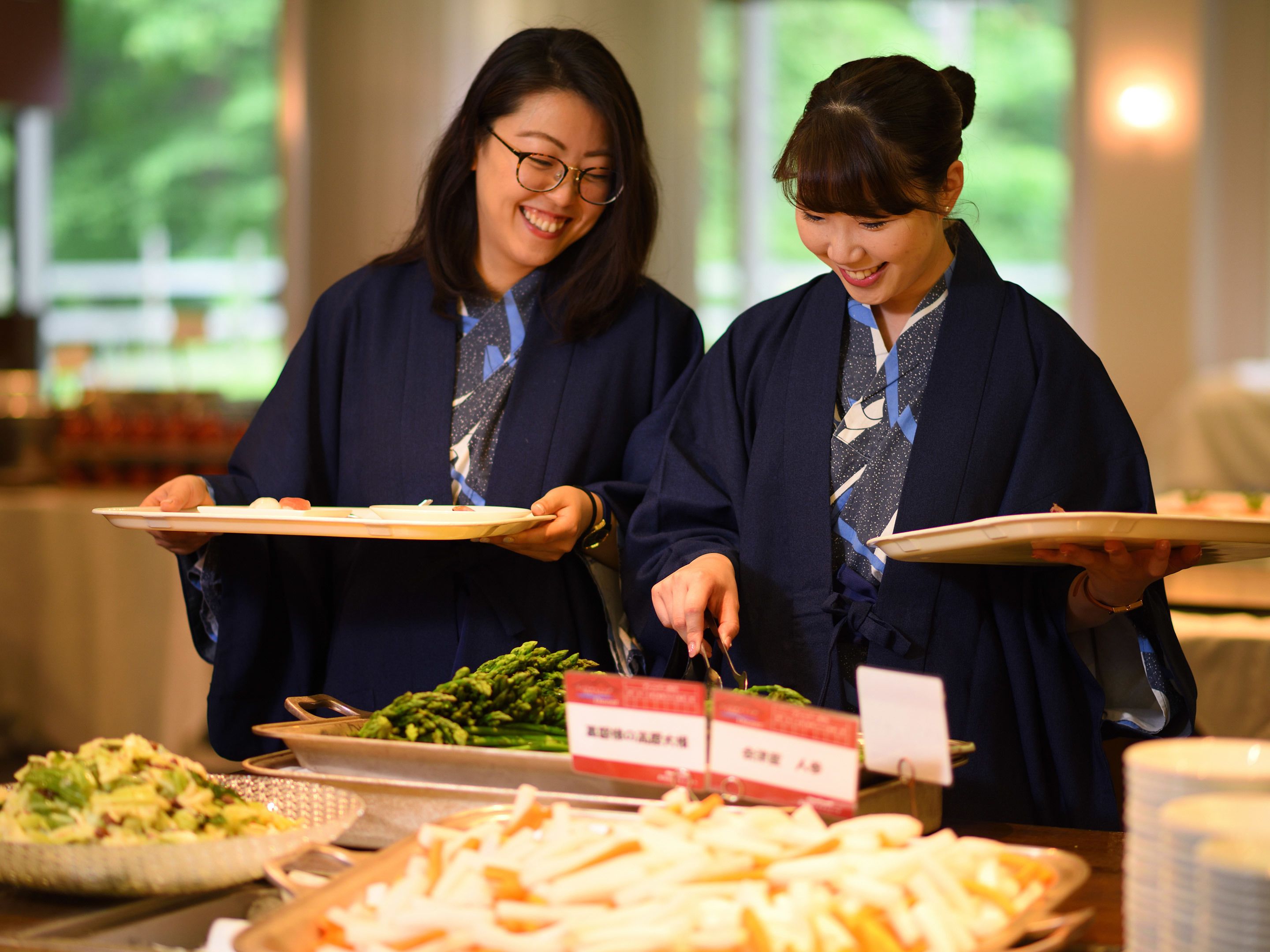 Zwei Personen in traditioneller Kleidung servieren Speisen an einem Buffet mit verschiedenen Gerichten und einem Schild, das den Preis angibt.