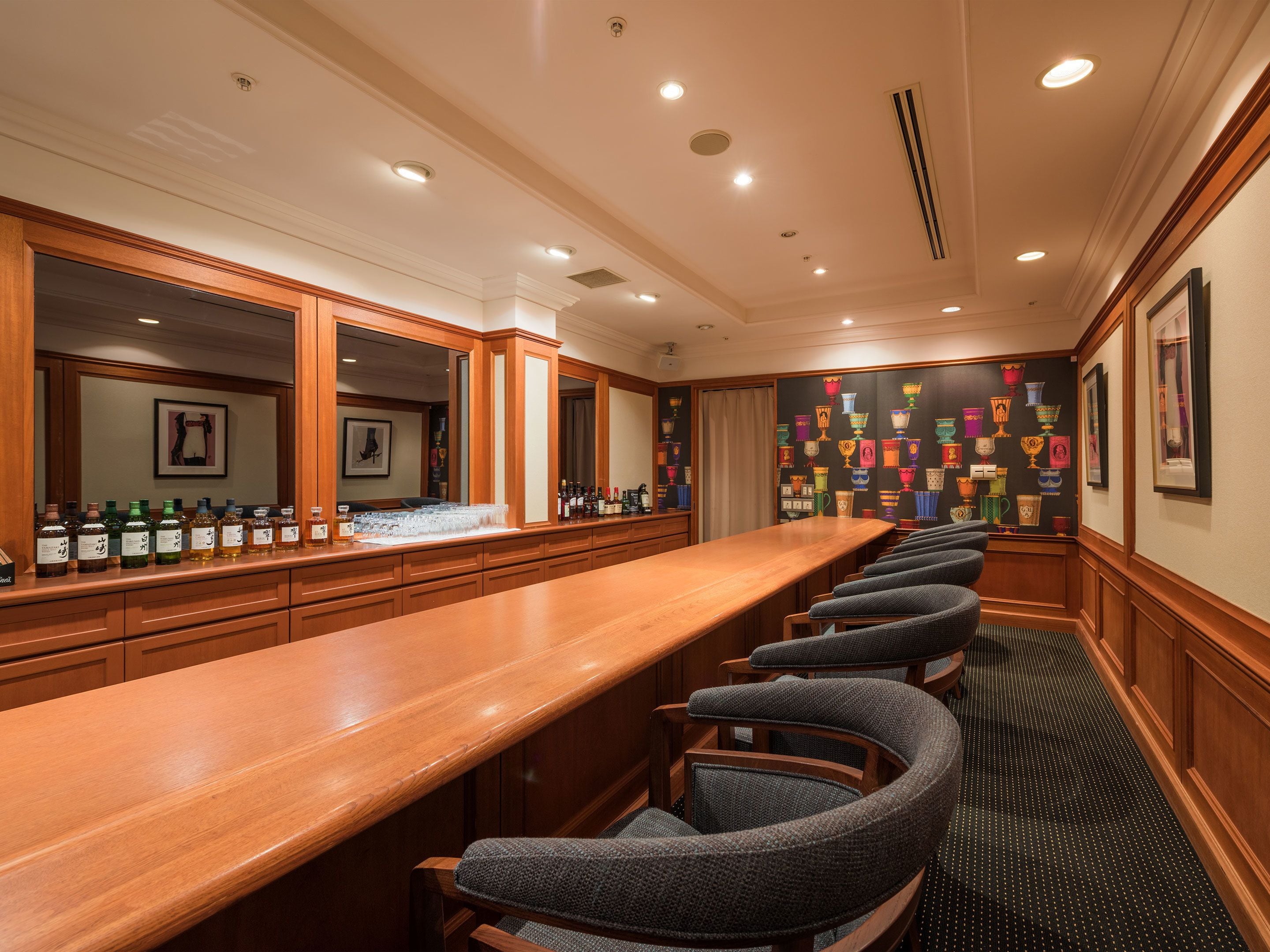 A bar area with a long wooden counter, multiple bar stools, and a wall decorated with colorful ceramic mugs.