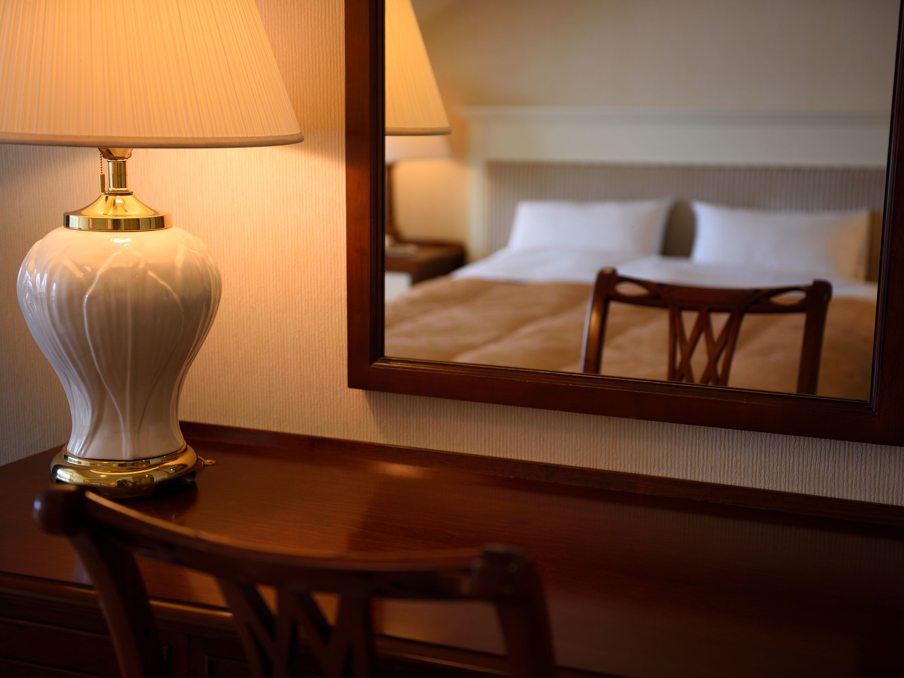A hotel room with a bedside lamp, a wooden chair, and a mirror reflecting a bed and nightstand.