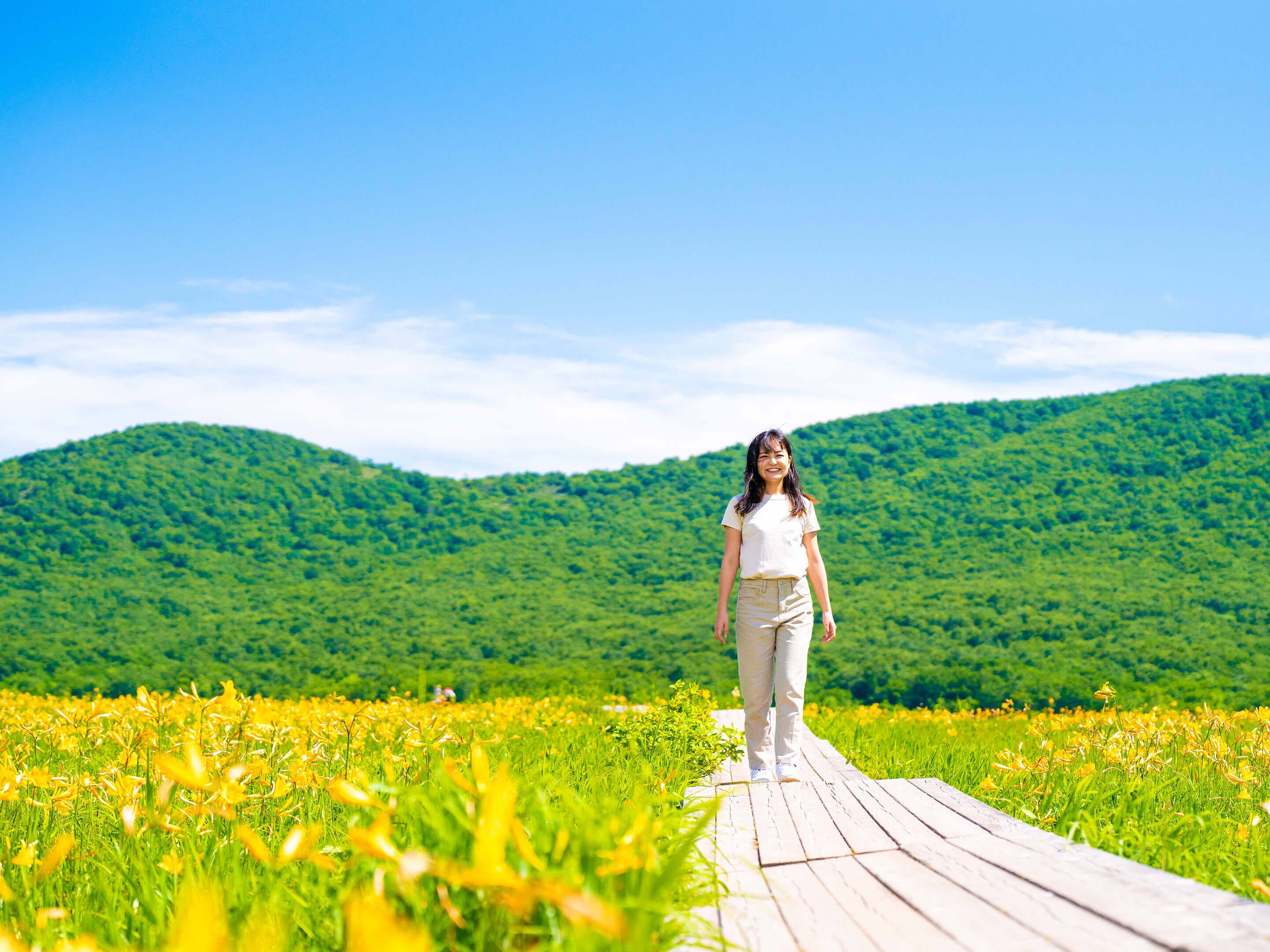 A person walking on a wooden boardwalk through a field of yellow flowers with green hills in the background.