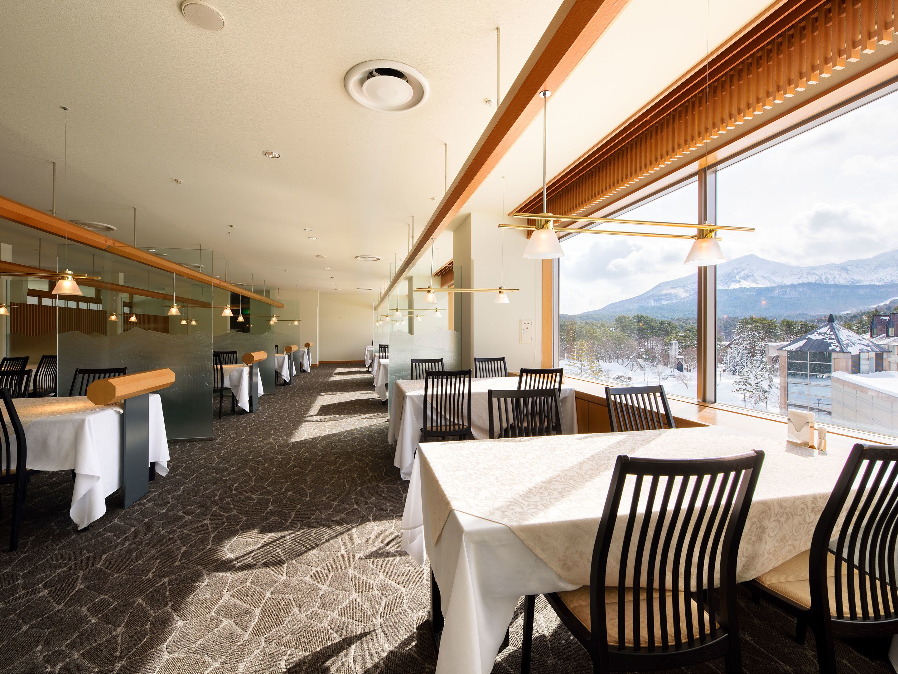 A restaurant with a long table layout, large windows with a mountain view, and modern pendant lighting.