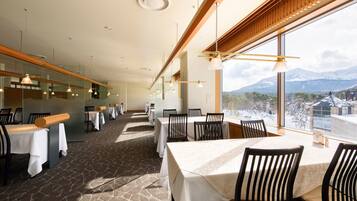 A restaurant with a long table layout, large windows with a mountain view, and modern pendant lighting.