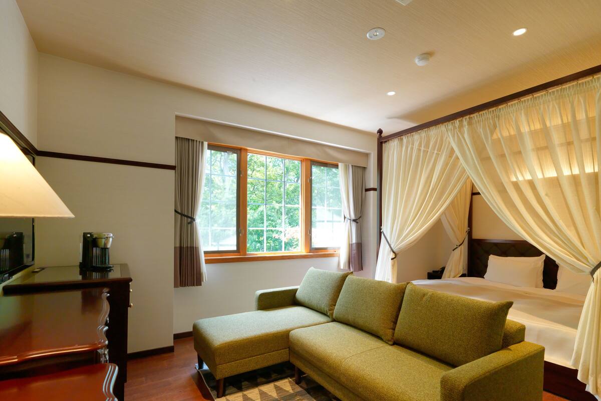 A hotel room with a sofa, a bed with a canopy, a desk with a lamp, and a window with curtains.