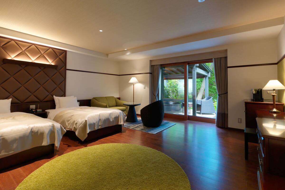 A hotel room with two beds, a green chair, a small table with a lamp, and a view of the outdoors through a sliding glass door.