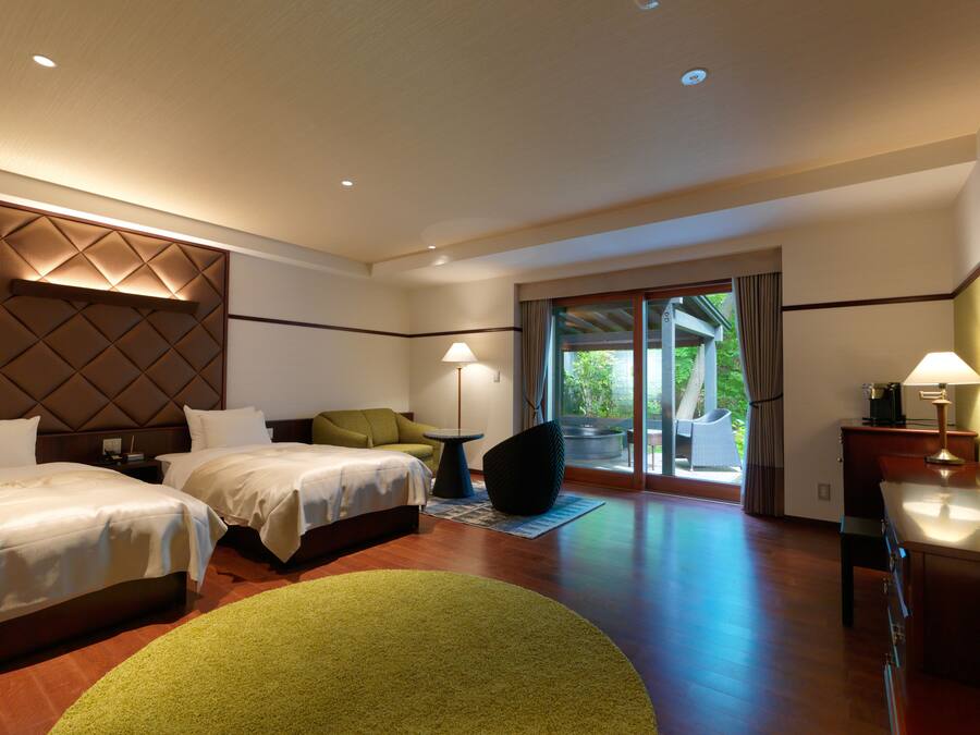 A hotel room with two beds, a green chair, a small table with a lamp, and a view of the outdoors through a sliding glass door.