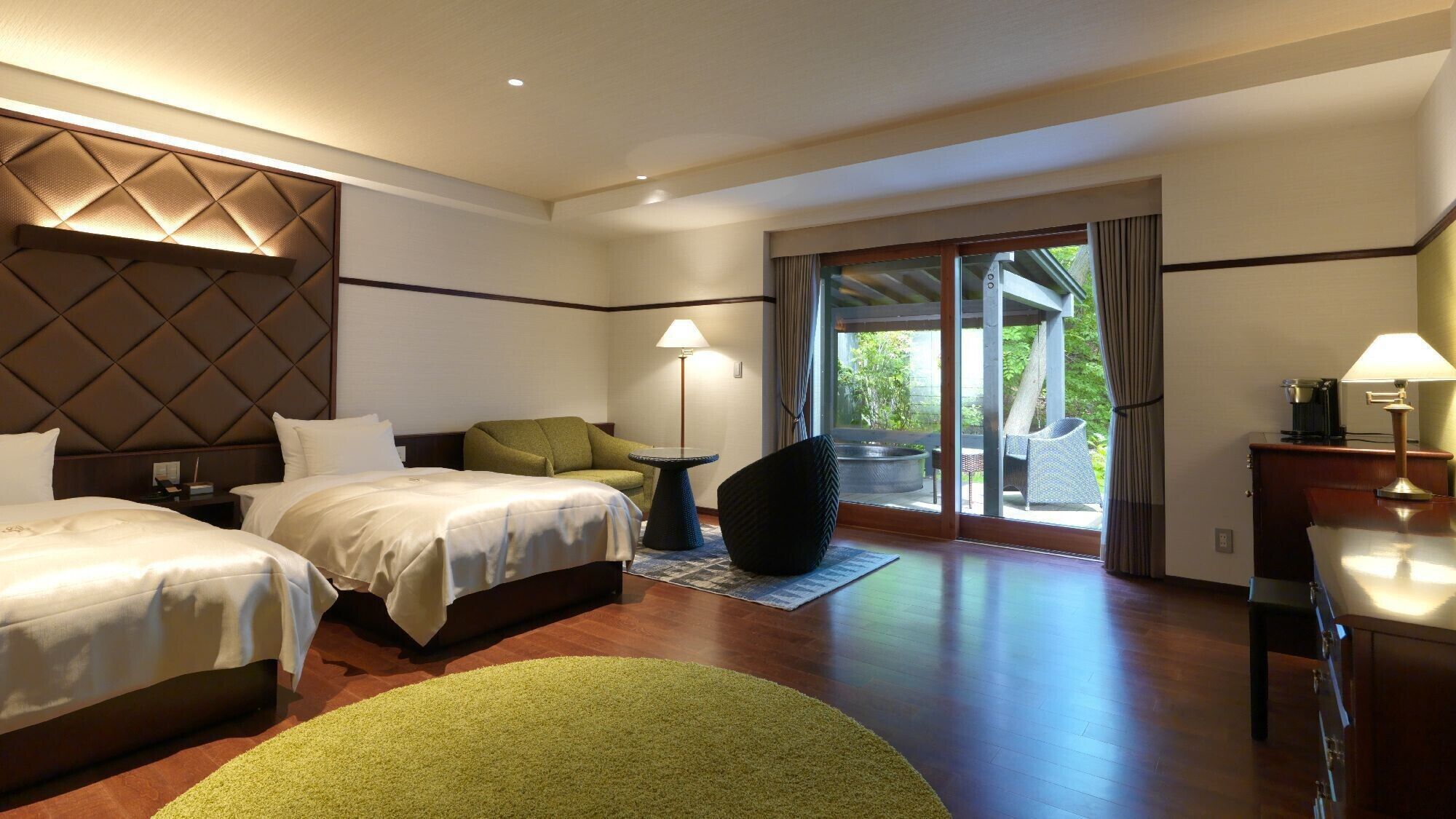A hotel room with two beds, a sofa, a small table, a lamp, and a view of the outdoors.