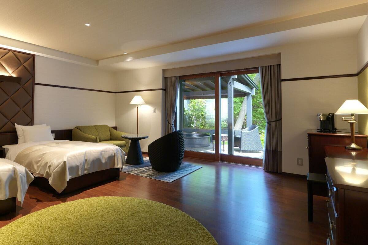 A hotel room with two beds, a sofa, a small table, a lamp, and a view of the outdoors.